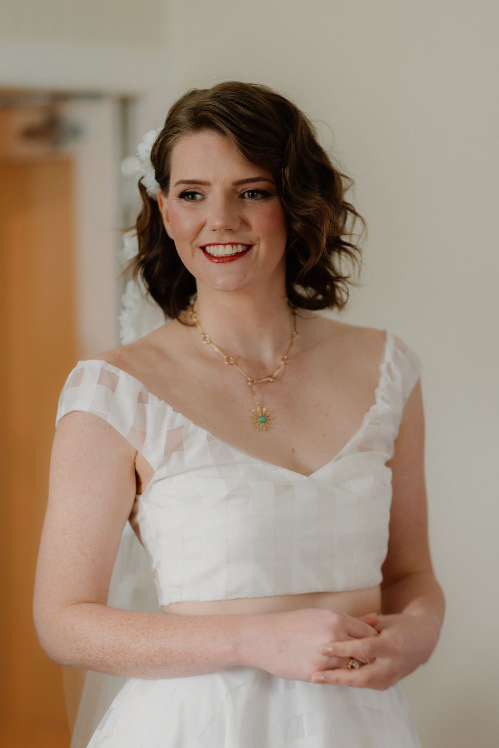 Bride in white off-shoulder wedding dress wearing a green pendant necklace, smiling warmly.