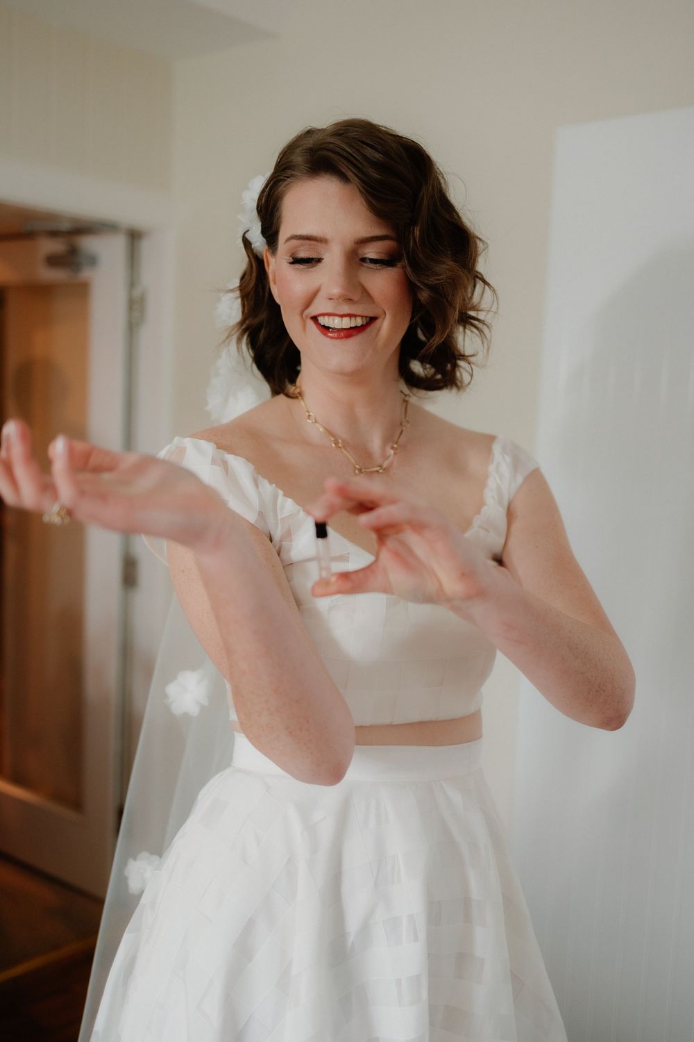 Smiling bride in white two-piece wedding dress applying perfume, wearing veil and gold necklace.