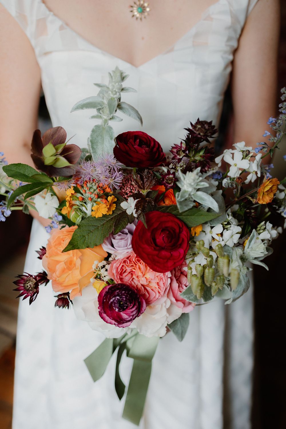 Bride holding a lush wedding bouquet with red ranunculus, peach roses, and wildflowers tied with sage green ribbon.