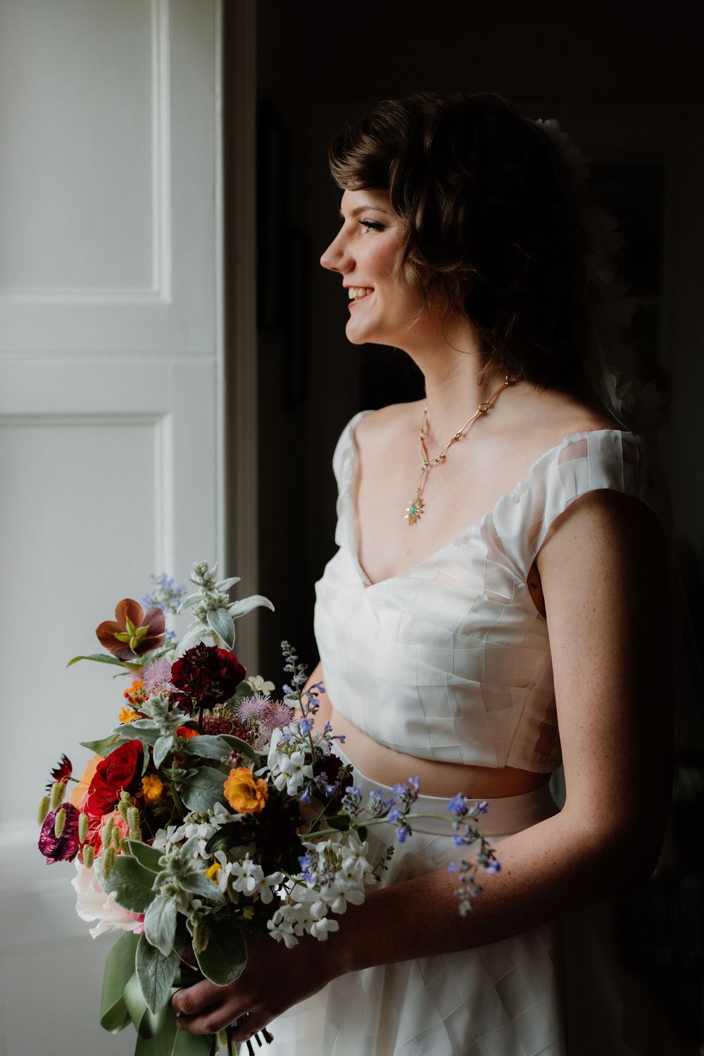 Smiling bride in white crop top gown holds colorful wildflower bouquet while standing by a window in soft natural light.