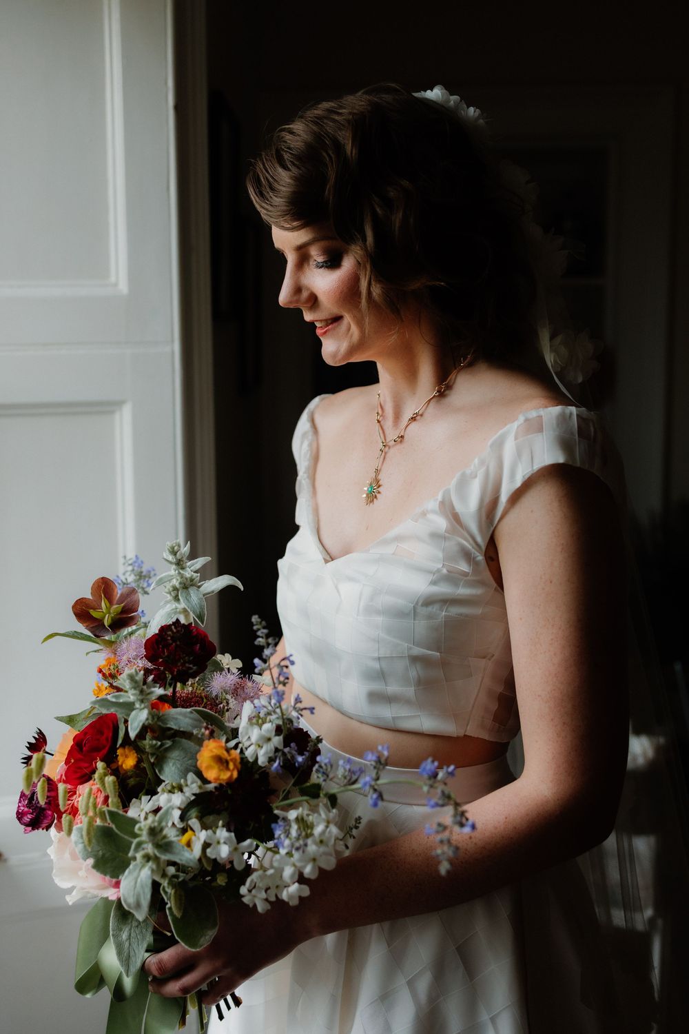 Smiling bride in white two-piece gown holding a colorful wildflower bouquet, wearing a gold necklace near a window.
