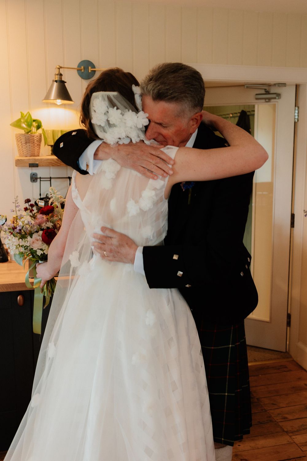 Bride in white gown and veil shares emotional hug with man in Scottish kilt at wedding venue.