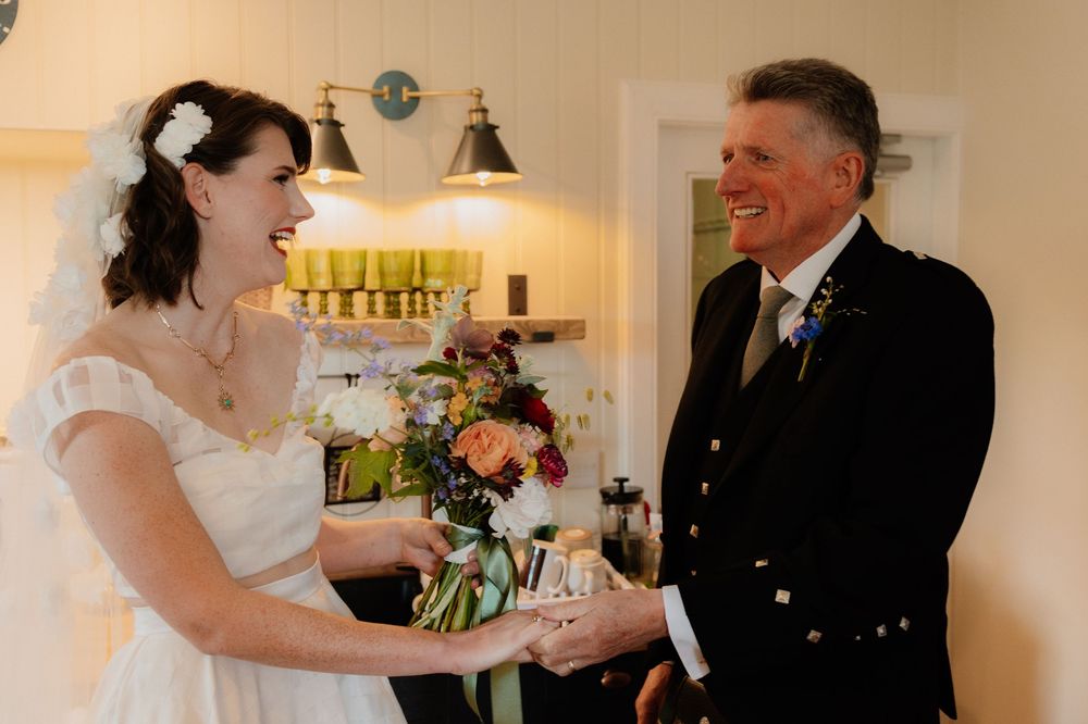 Bride in white lace gown holding colorful bouquet laughing with man in Scottish kilt during intimate wedding ceremony indoors.