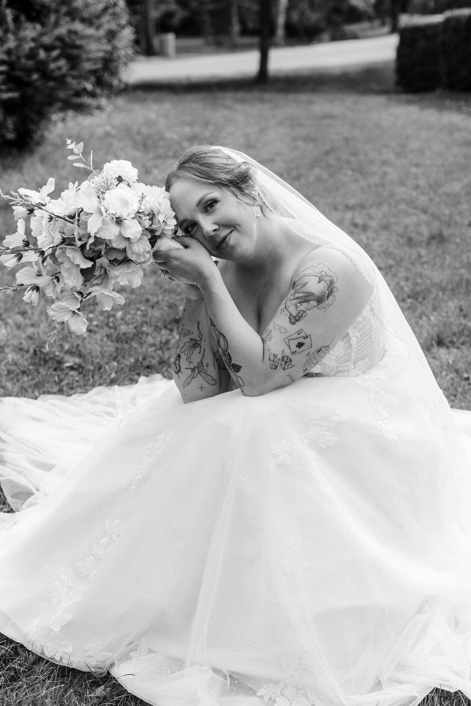 Black and white photo of a smiling bride in a tulle gown smelling her floral bouquet while seated outdoors.