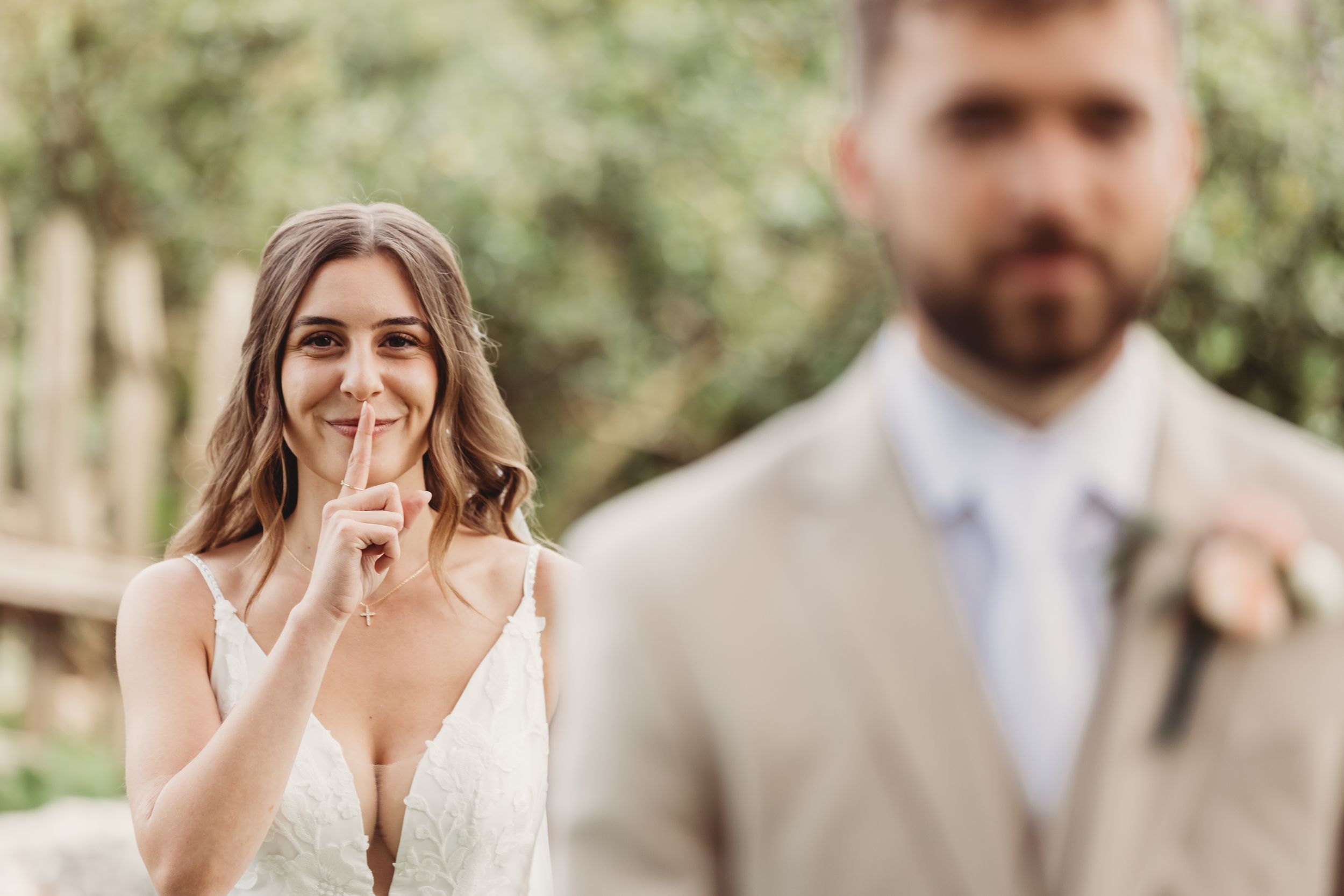 Bride in white lace gown smiling with finger to lips during wedding ceremony, groom in tan suit in foreground.