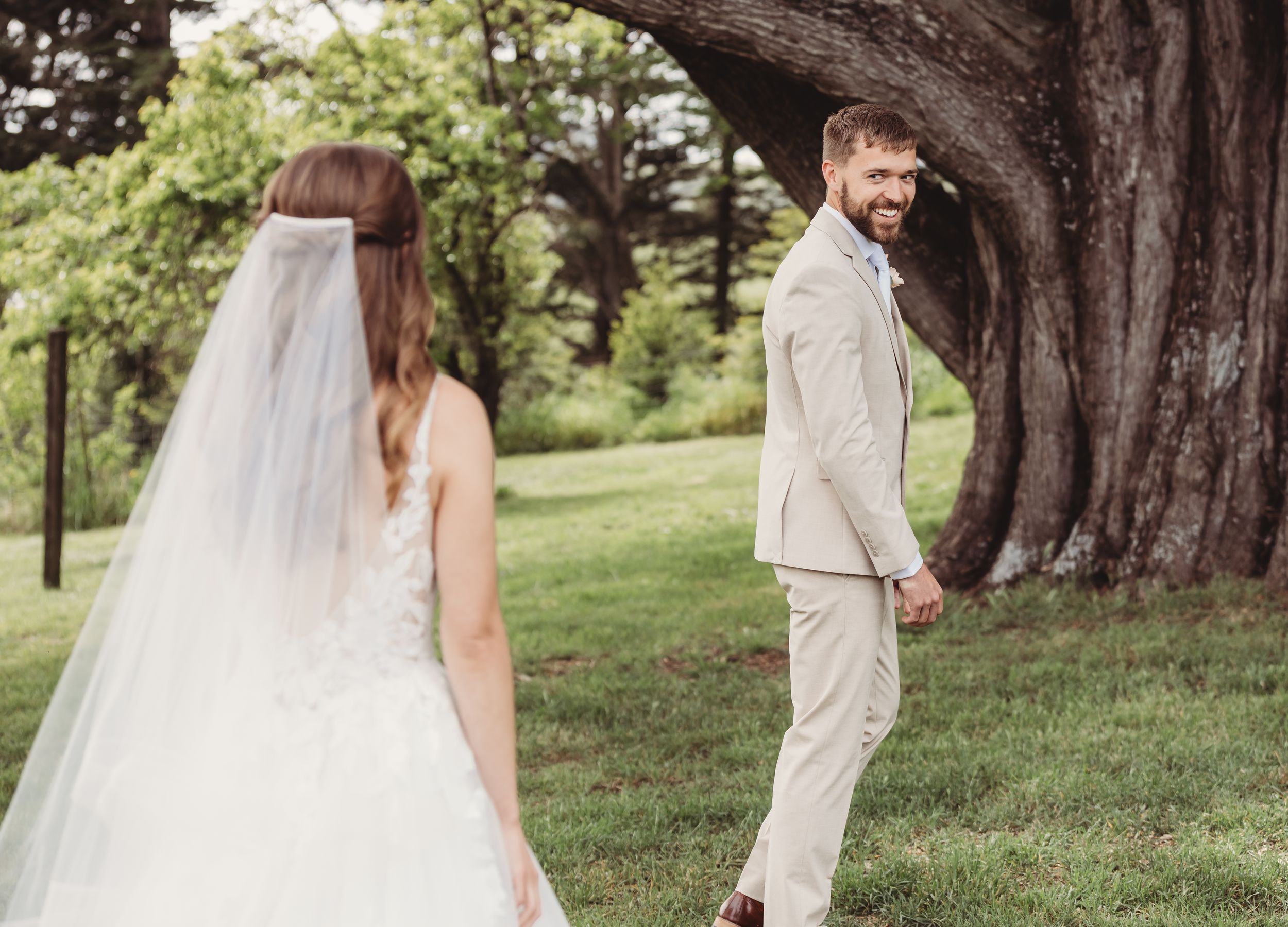 Groom in tan suit smiling at bride in white wedding gown and veil outdoors near a large tree.