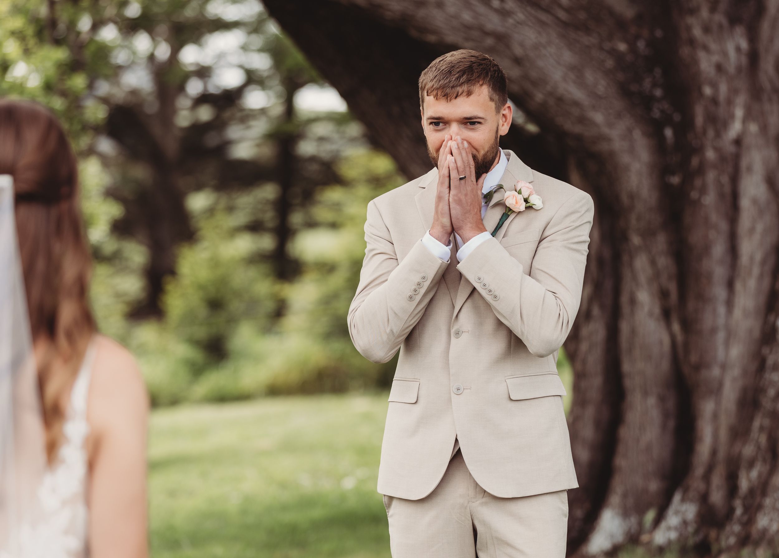 Emotional groom in beige suit with hands clasped to face seeing bride for first time at outdoor wedding ceremony.