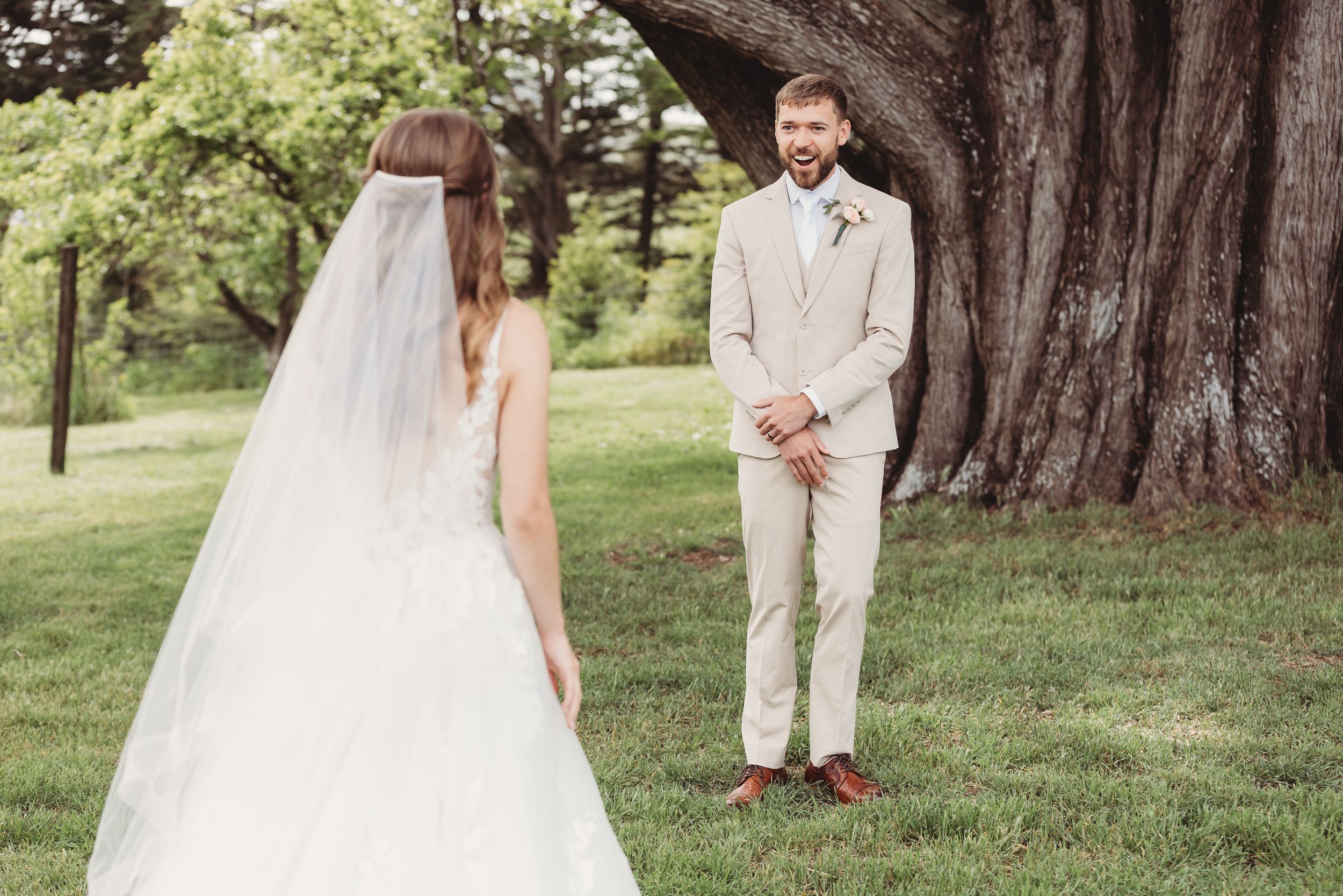 Groom in beige suit reacts emotionally seeing bride in white wedding gown for first look outdoors.