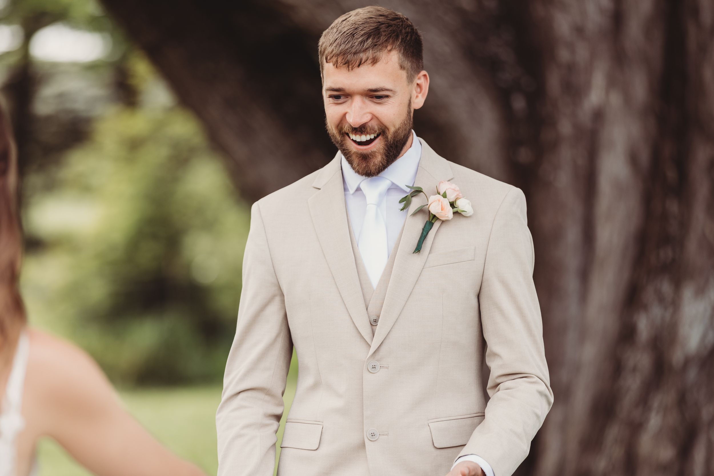 Smiling groom in beige suit with boutonniere standing outdoors at wedding ceremony.