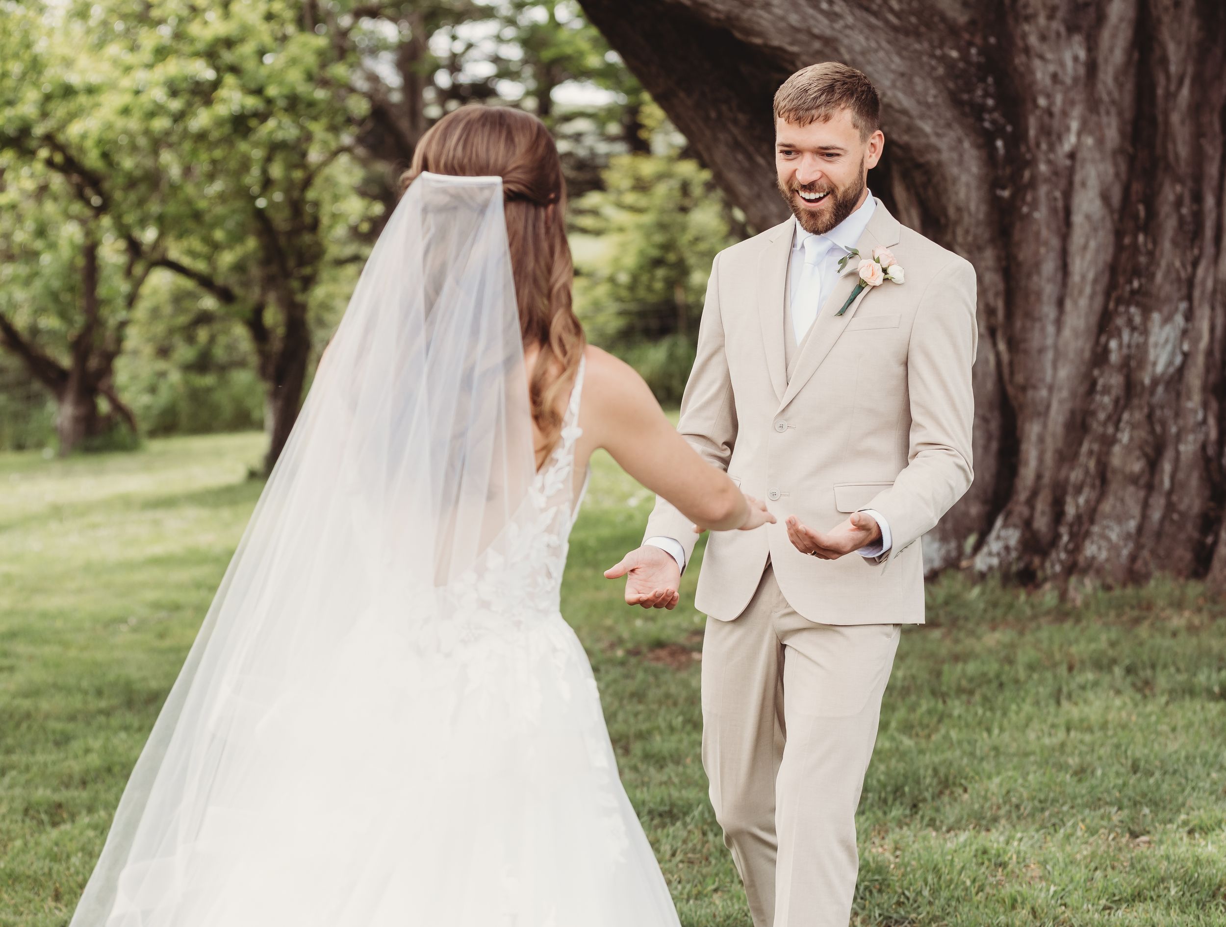 A groom in a beige suit smiles at his bride in a white wedding dress and veil during an outdoor first look.