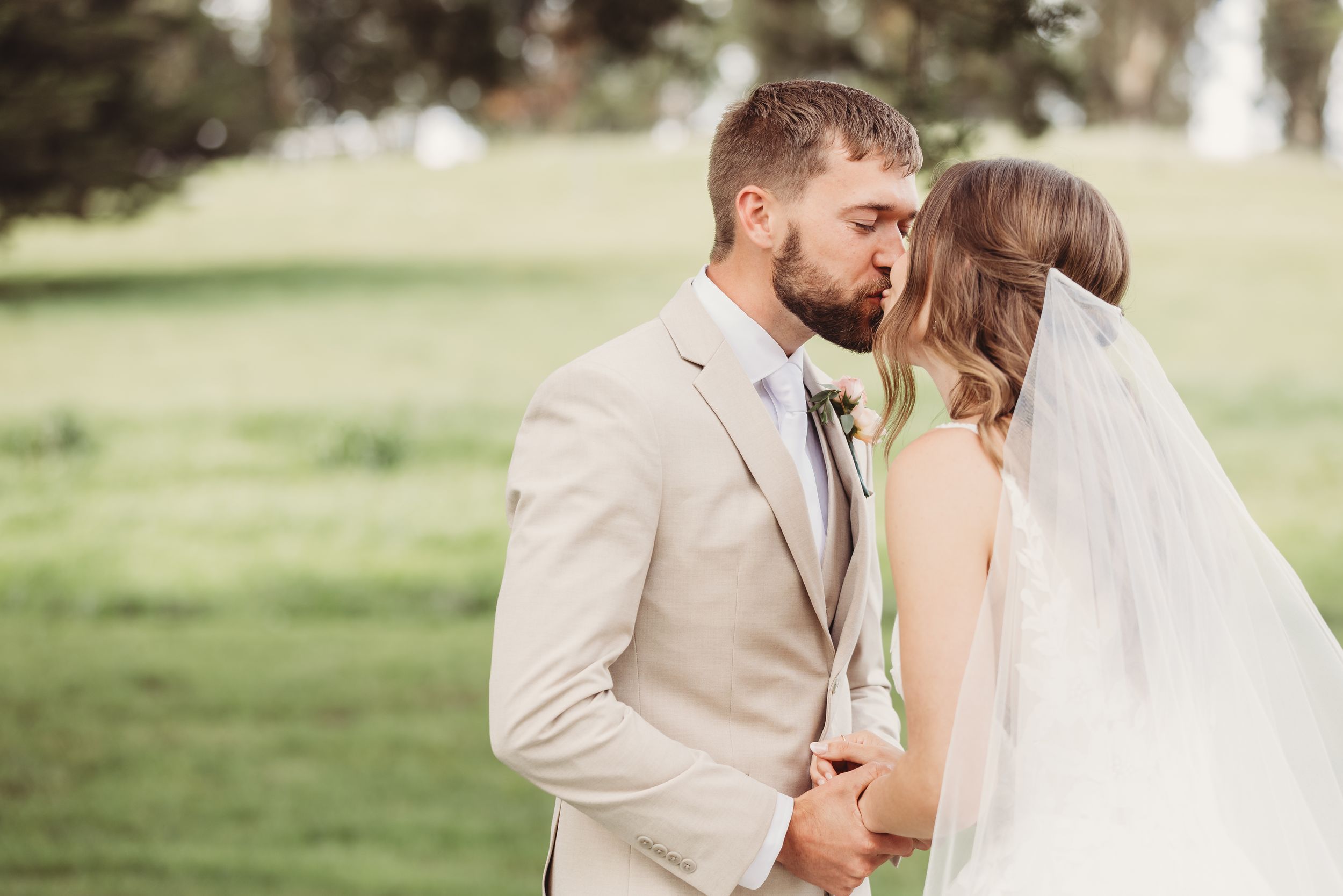 A bride and groom share a romantic kiss outdoors on their wedding day, surrounded by lush green grass.