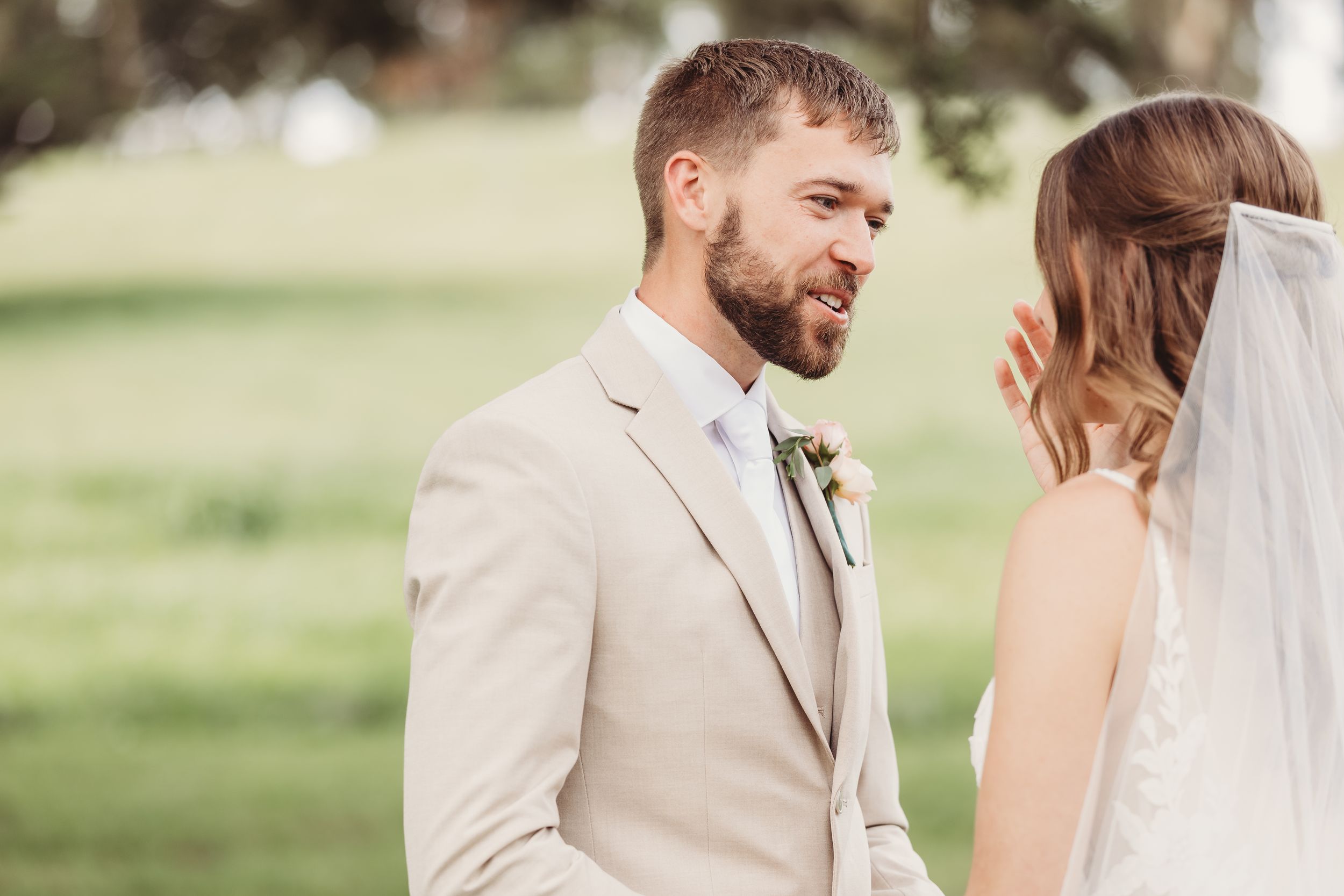 Groom in beige suit smiling at bride in white veil during outdoor wedding ceremony on green lawn.