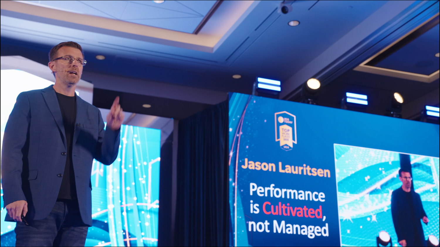 Jason Lauritsen speaks on stage at a conference, with a slide reading Performance is Cultivated, not Managed.
