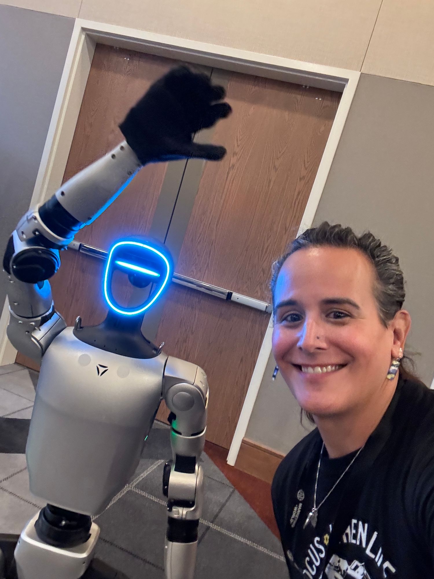 A woman smiles beside a humanoid robot with glowing blue face display raising its hand indoors.