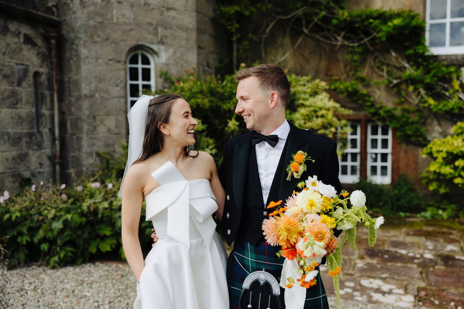 Bride in white strapless gown and groom in kilt share a joyful moment holding a colourful bouquet in front of Glasgow wedding Venue