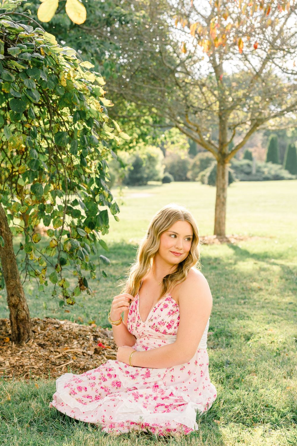 Young woman in floral pink dress sitting on grass beside a tree in a sunny green park.