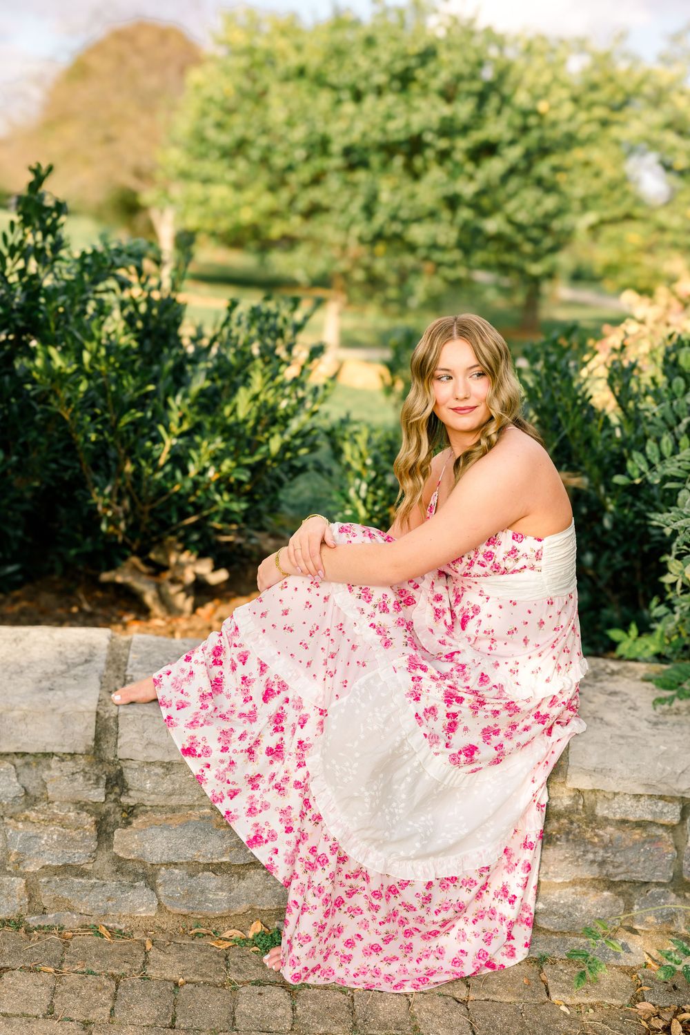 Young woman in pink floral maxi dress sitting on stone wall surrounded by lush green shrubs outdoors.