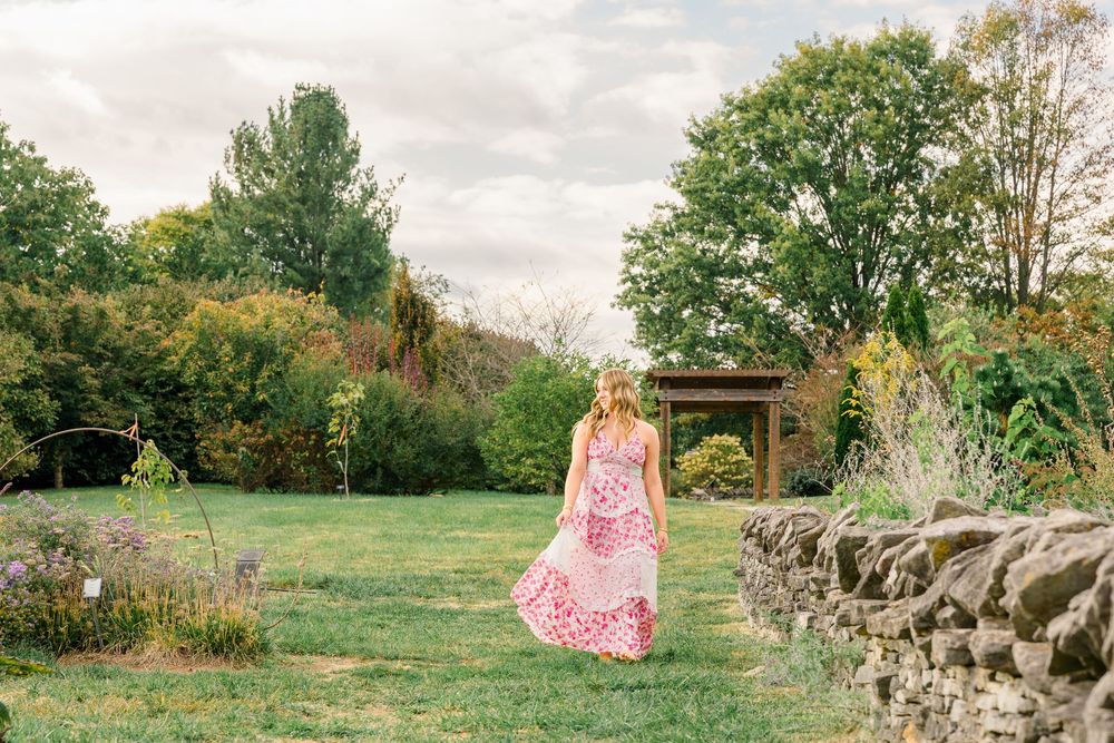Woman in pink floral maxi dress walking through lush green garden with stone wall and pergola.