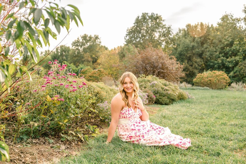 Young blonde woman in floral dress sitting on grass beside blooming pink flowers in a lush green garden.