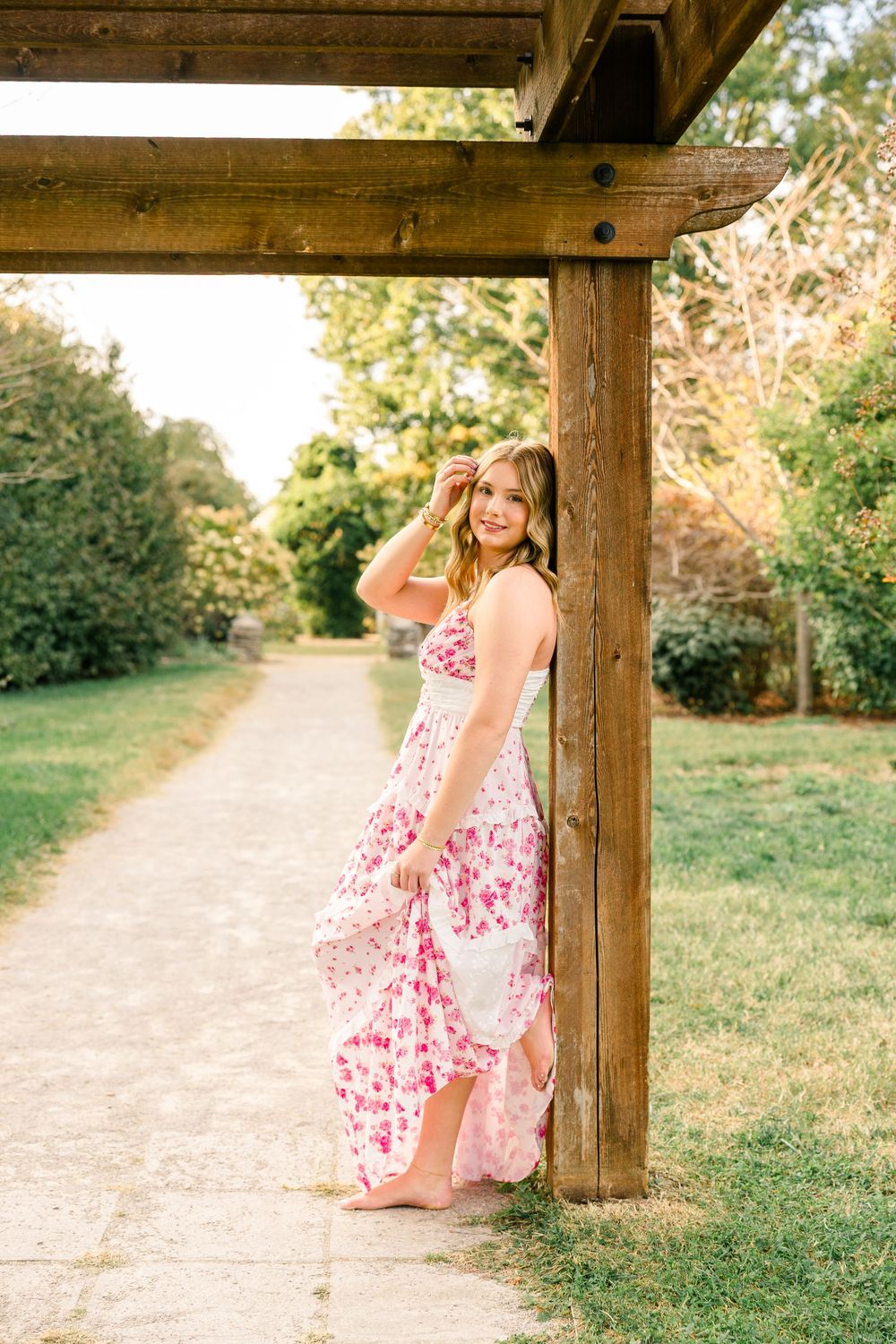 Young woman in pink floral maxi dress poses by wooden pergola on garden path surrounded by lush greenery.