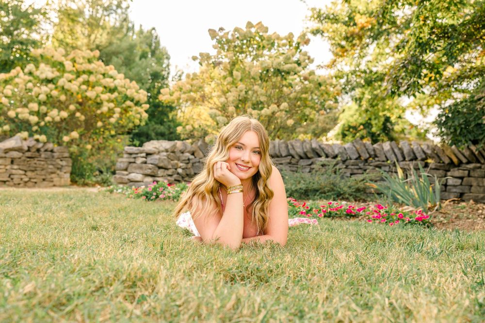 Young blonde woman lying on green grass in a sunny garden with stone wall and colorful flowers in background.