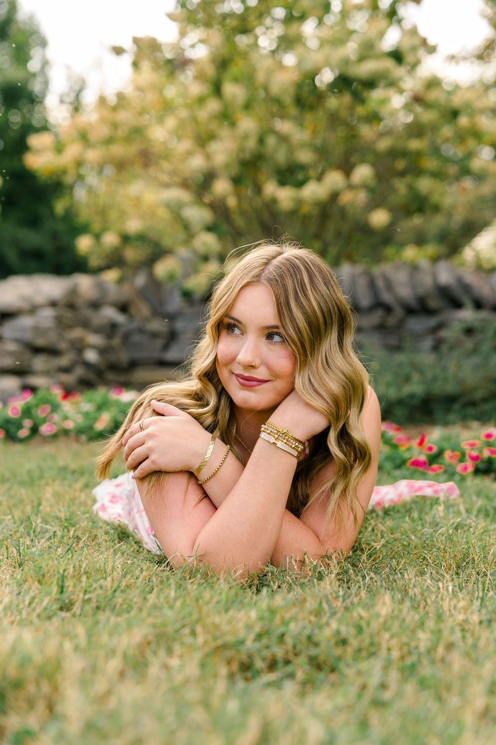 Young woman with wavy blonde hair lying on green grass, wearing gold bracelets, with colorful garden flowers in background.
