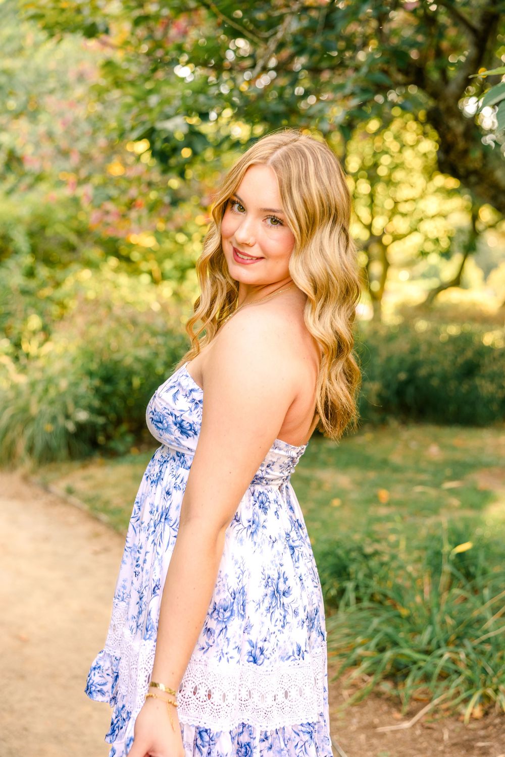 Young blonde woman in blue floral sundress smiling outdoors in lush green garden setting.