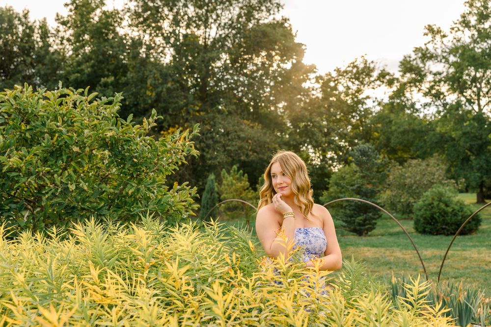 Young woman in floral dress poses among golden wildflowers at sunset in a lush green garden.