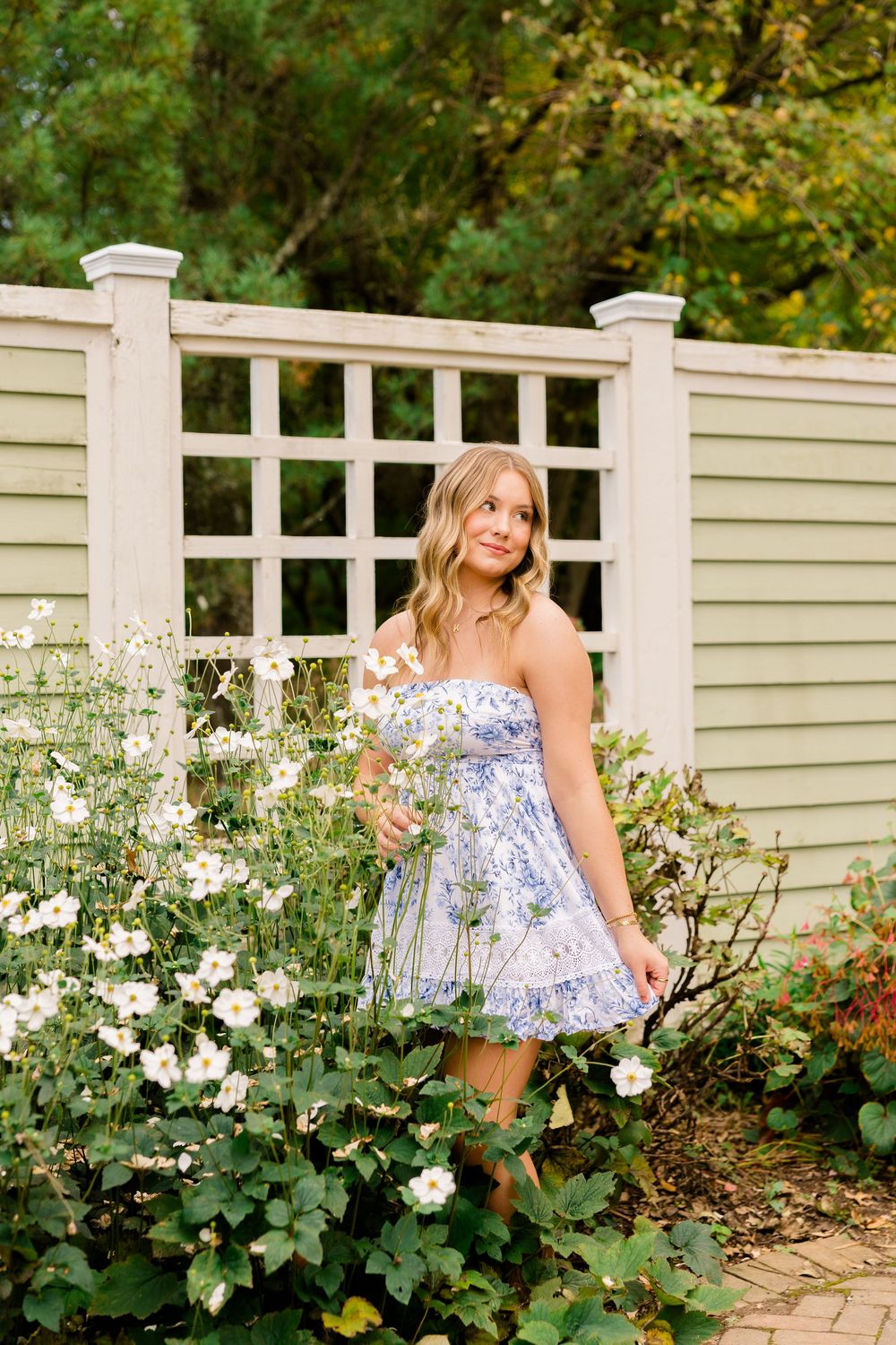A young woman in a blue floral strapless dress poses among white flowers beside a white garden trellis fence.