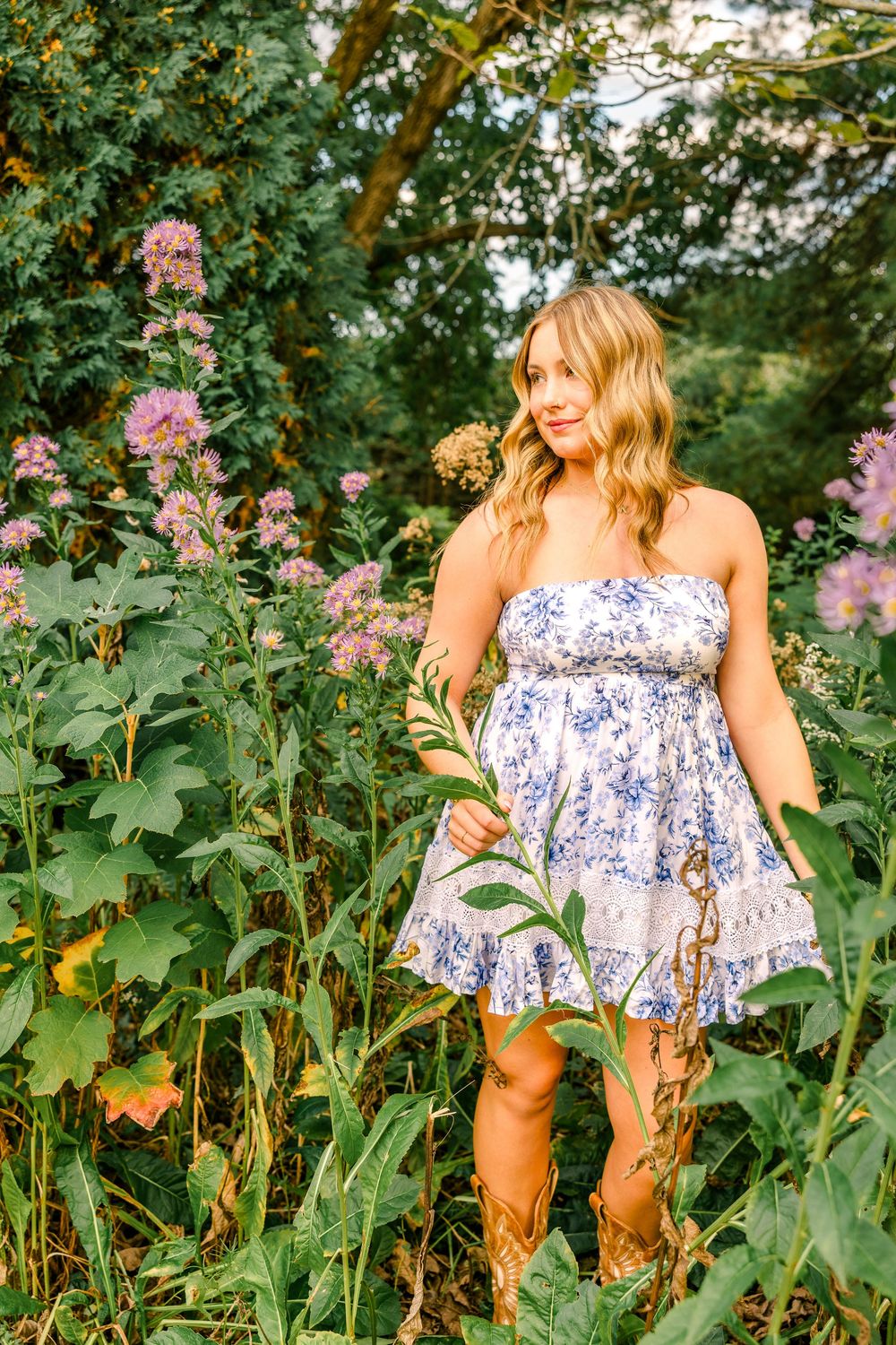 Young blonde woman in floral strapless mini dress and cowboy boots standing among wildflowers in a lush garden.