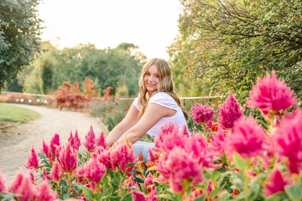 Young blonde woman smiling among vibrant pink celosia flowers in a sunny garden path setting.