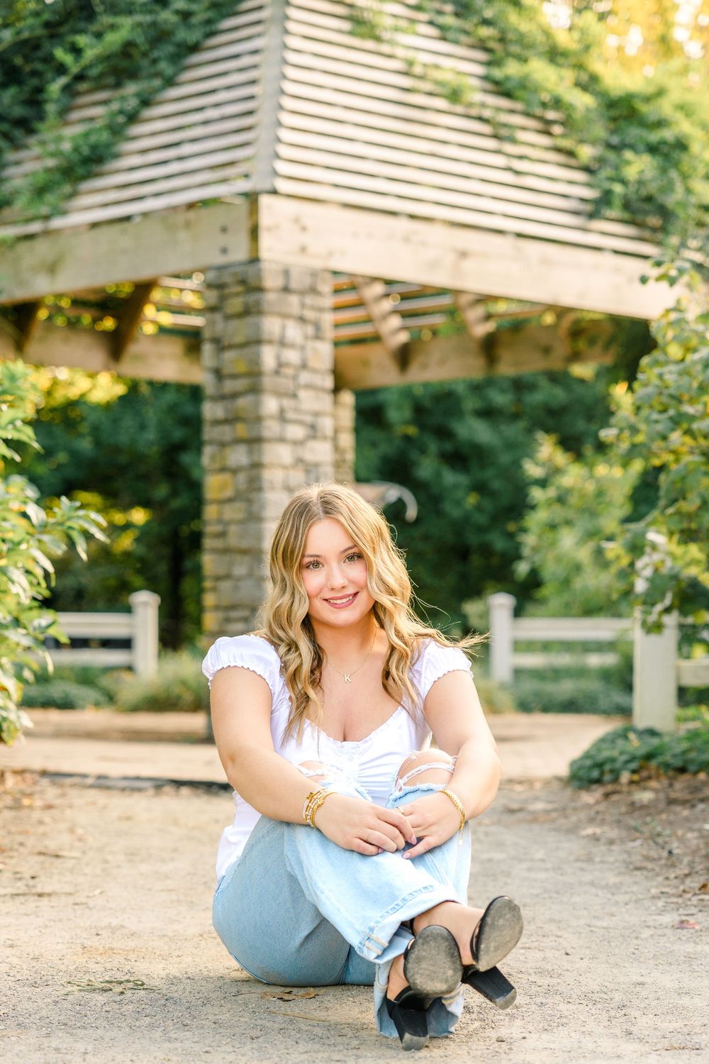 Young blonde woman sitting on a gravel path in light jeans and white top, smiling in front of a wooden gazebo.