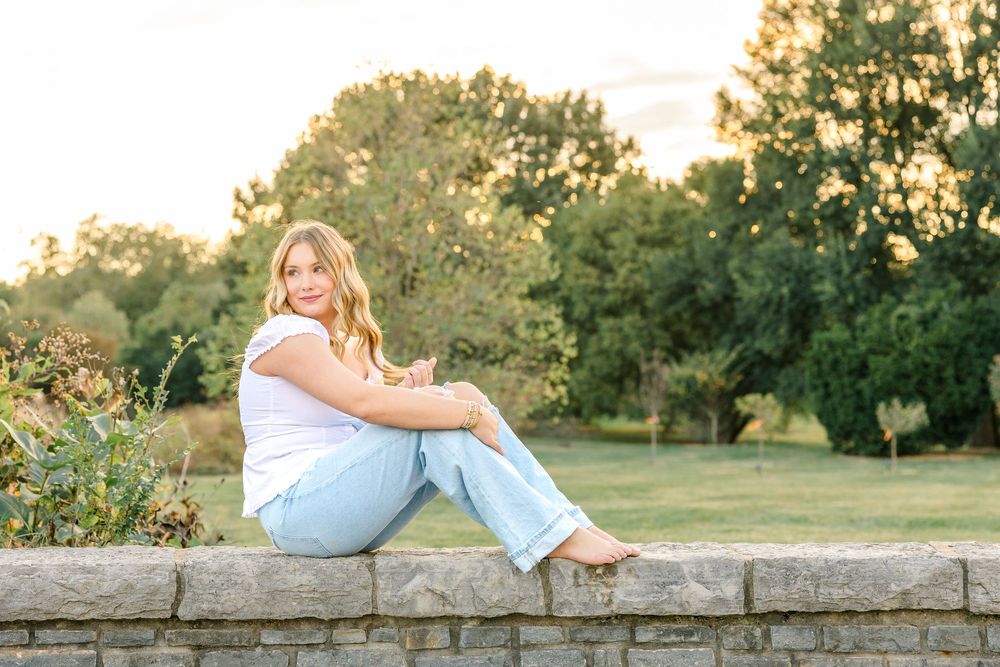 A young blonde woman sits on a stone wall in a park, wearing a white top and light blue jeans, smiling outdoors.