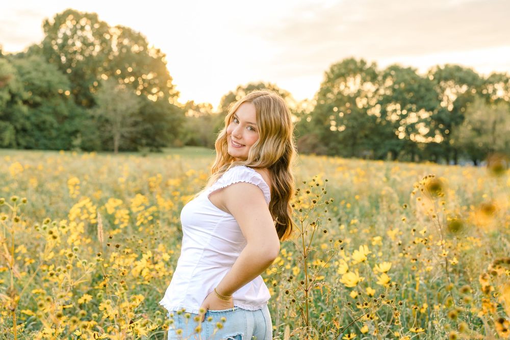 Young woman smiling in a sunflower field at golden hour, wearing a white top and denim shorts.