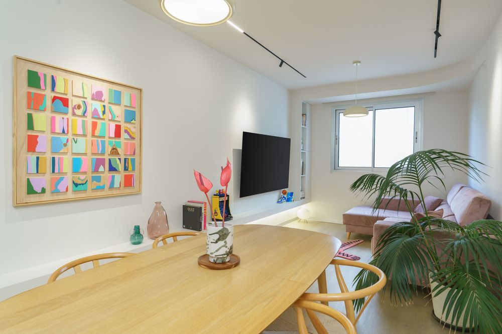 Modern living and dining room with colorful wall art, wooden table, TV, pink sofa, and tropical plant.