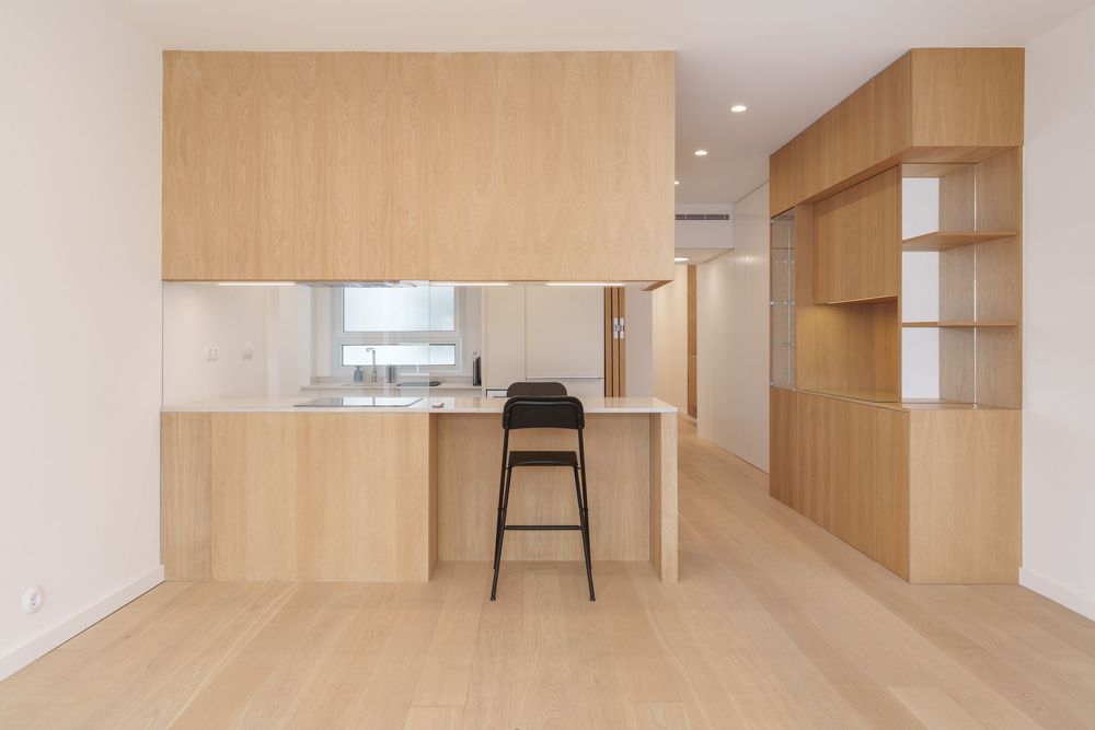 Modern minimalist kitchen with light wood cabinetry, island bar stool, and open shelving unit in a bright apartment.