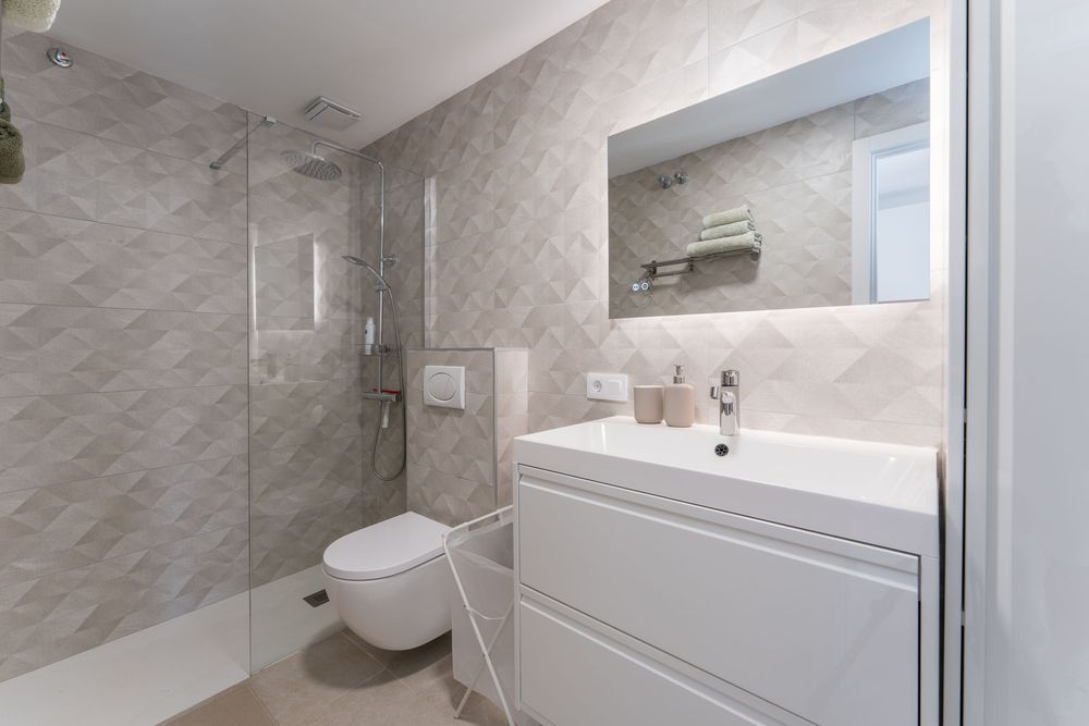 Modern white bathroom with walk-in glass shower, wall-hung toilet, geometric tiles, and large vanity mirror.