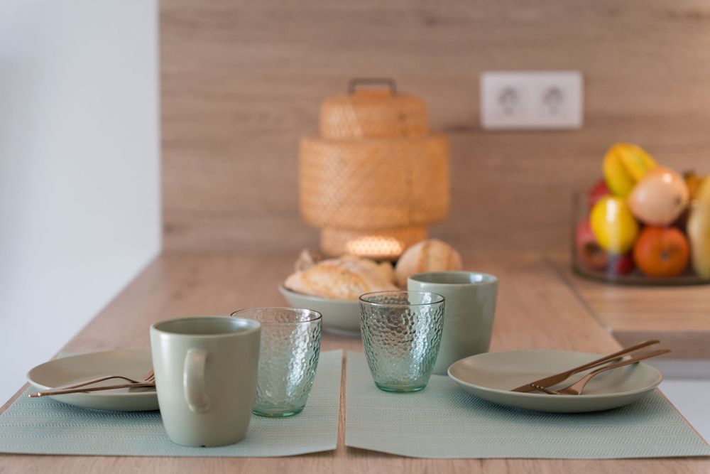 Minimalist kitchen table set with sage green cups, glasses, and plates on green placemats with bread and fruit in background.