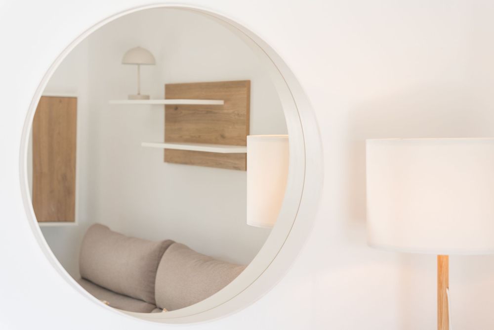 Round white mirror reflecting a minimalist room with wooden shelves, a small lamp, and a beige sofa with cushions.