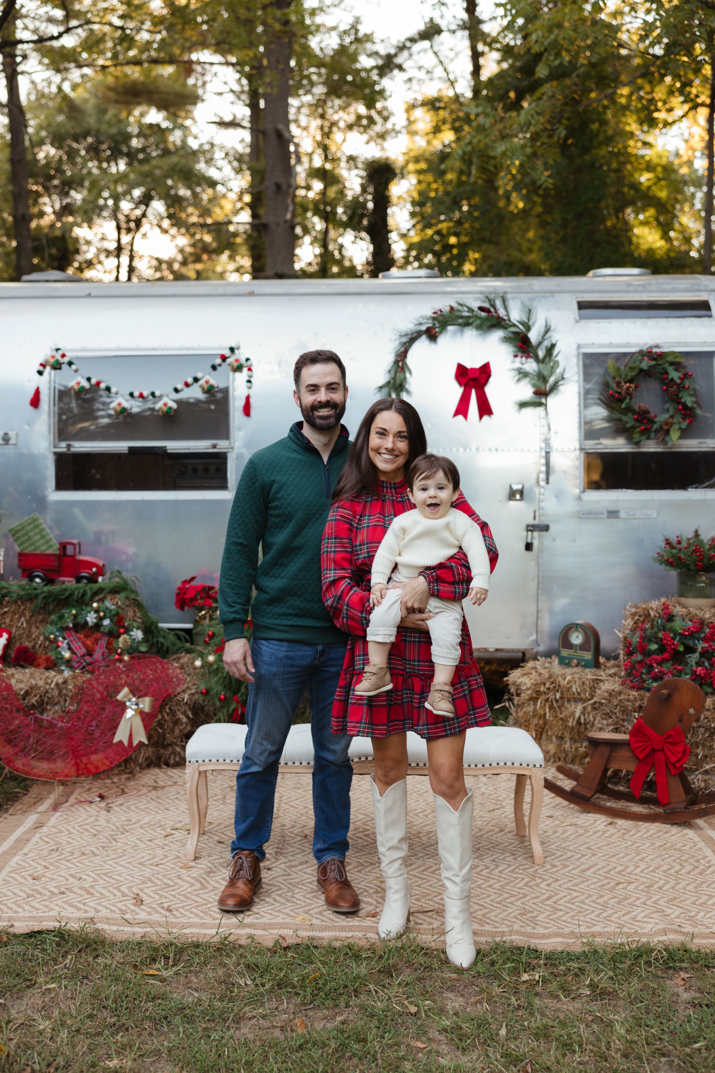 Family of three posing in front of a Christmas-decorated silver Airstream trailer surrounded by holiday wreaths and red bows.