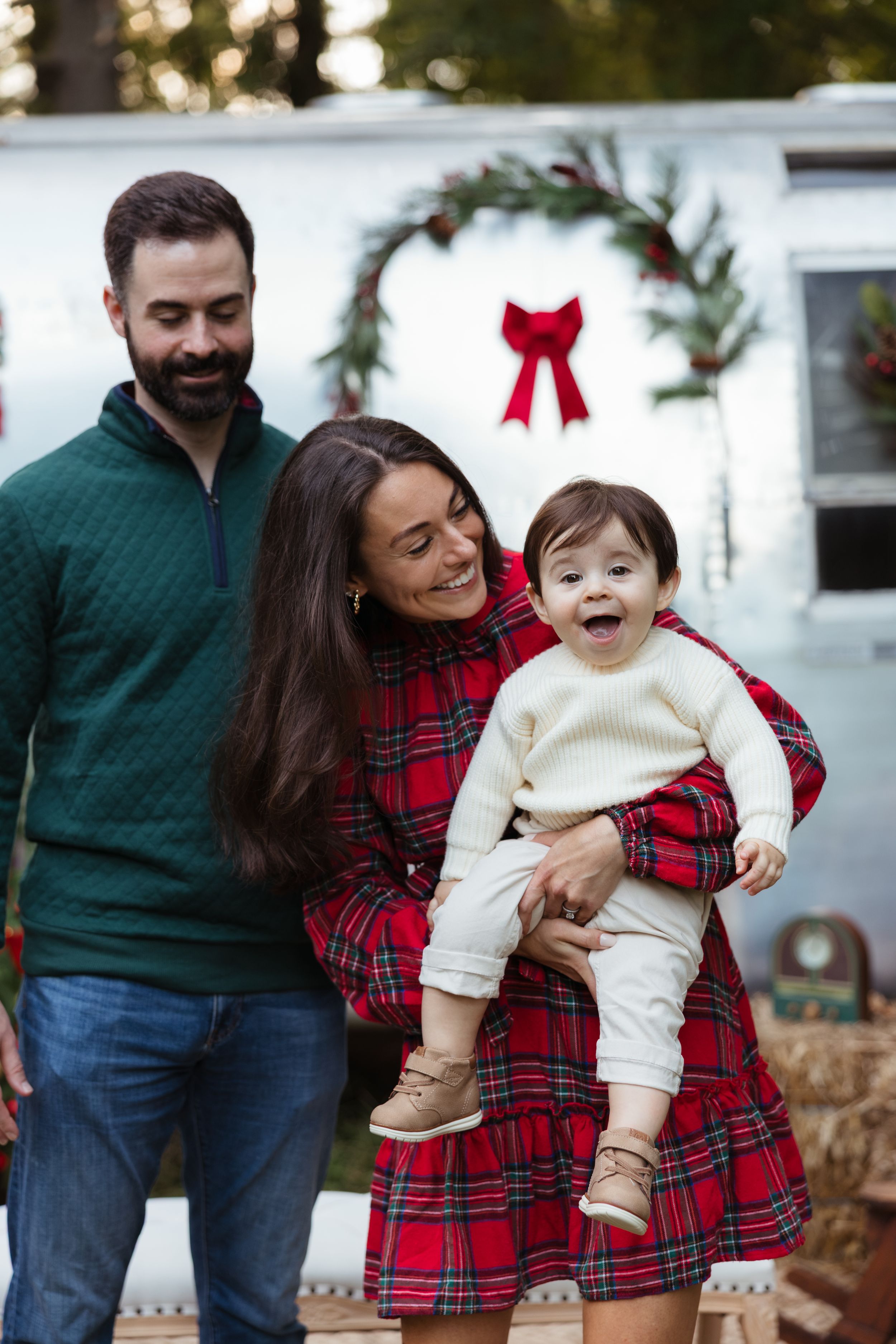 Happy family with baby in Christmas plaid outfits posing in front of a holiday-decorated trailer with wreath.