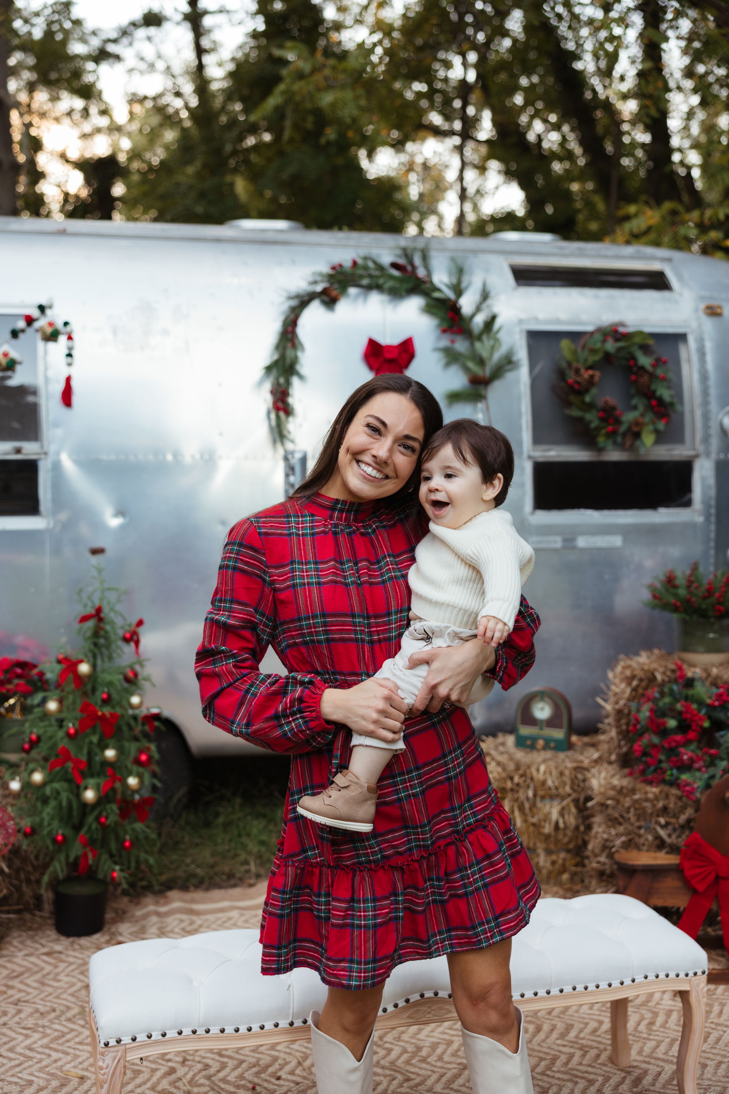 A smiling woman in a red plaid dress holds a baby near a decorated Airstream trailer with Christmas wreaths and holiday decor.