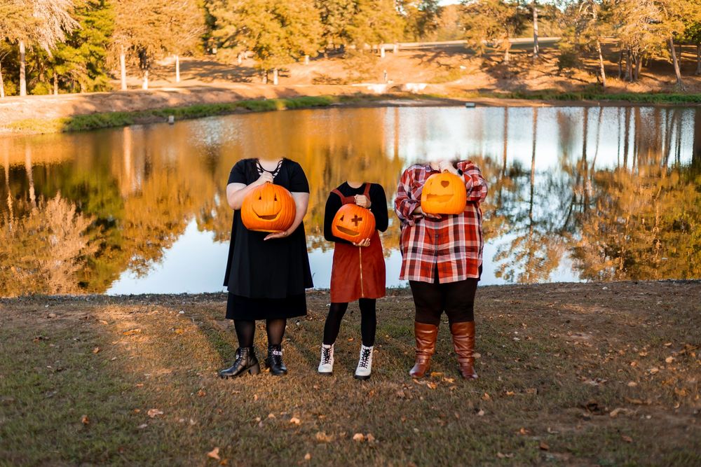 Three people holding orange pumpkins in front of their faces near a reflective pond during autumn.