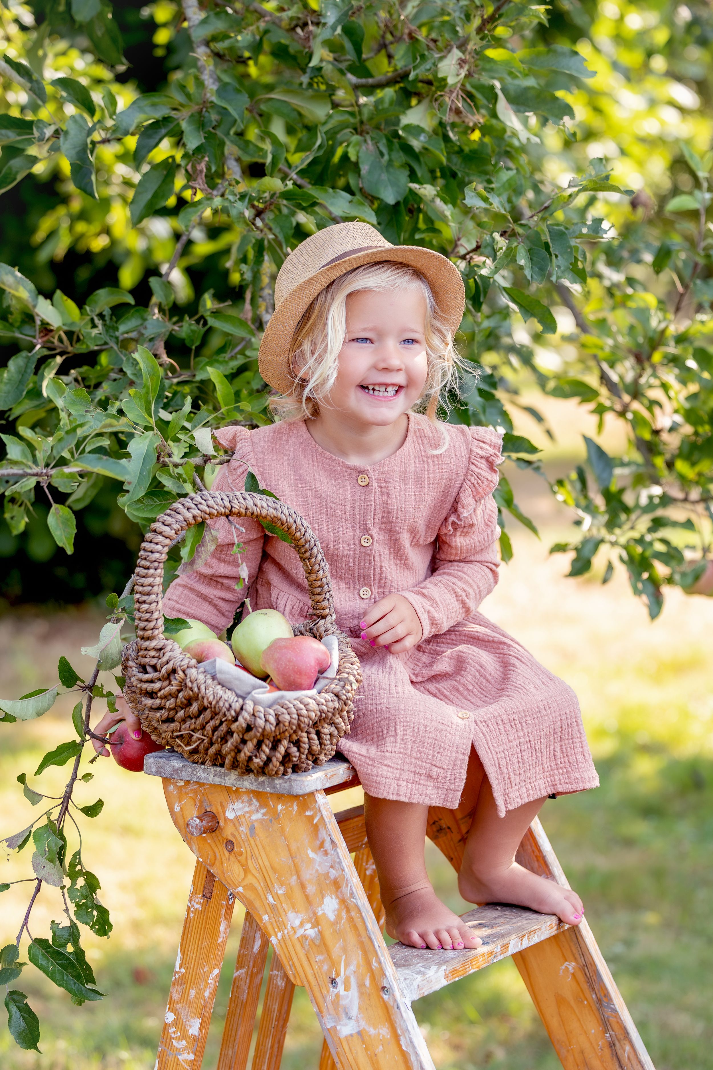meisje in roze jurk, zittend op een schilderstrap onder een appelboom. Op het krukje staat een rieten mandje gevuld met appels