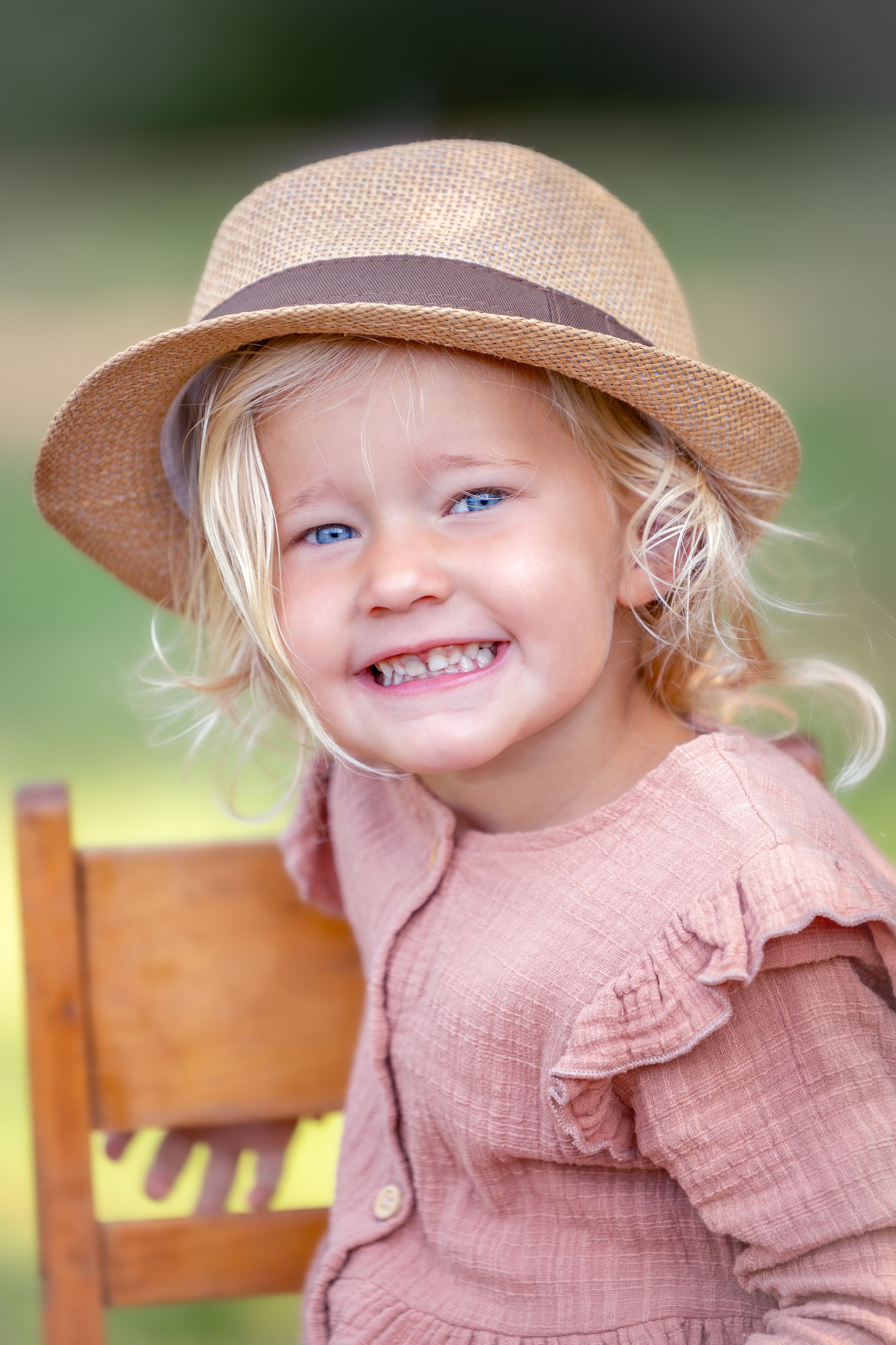 kinderportret van een meisje met blonde krullen. Ze draagt een strohoedje en zit op een houten kinderstoeltje.