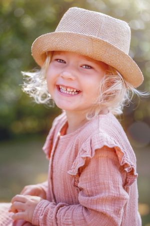 kinderportret van een meisje met blonde krullen. Lachend naar de camera met een strohoedje op haar hoofd.