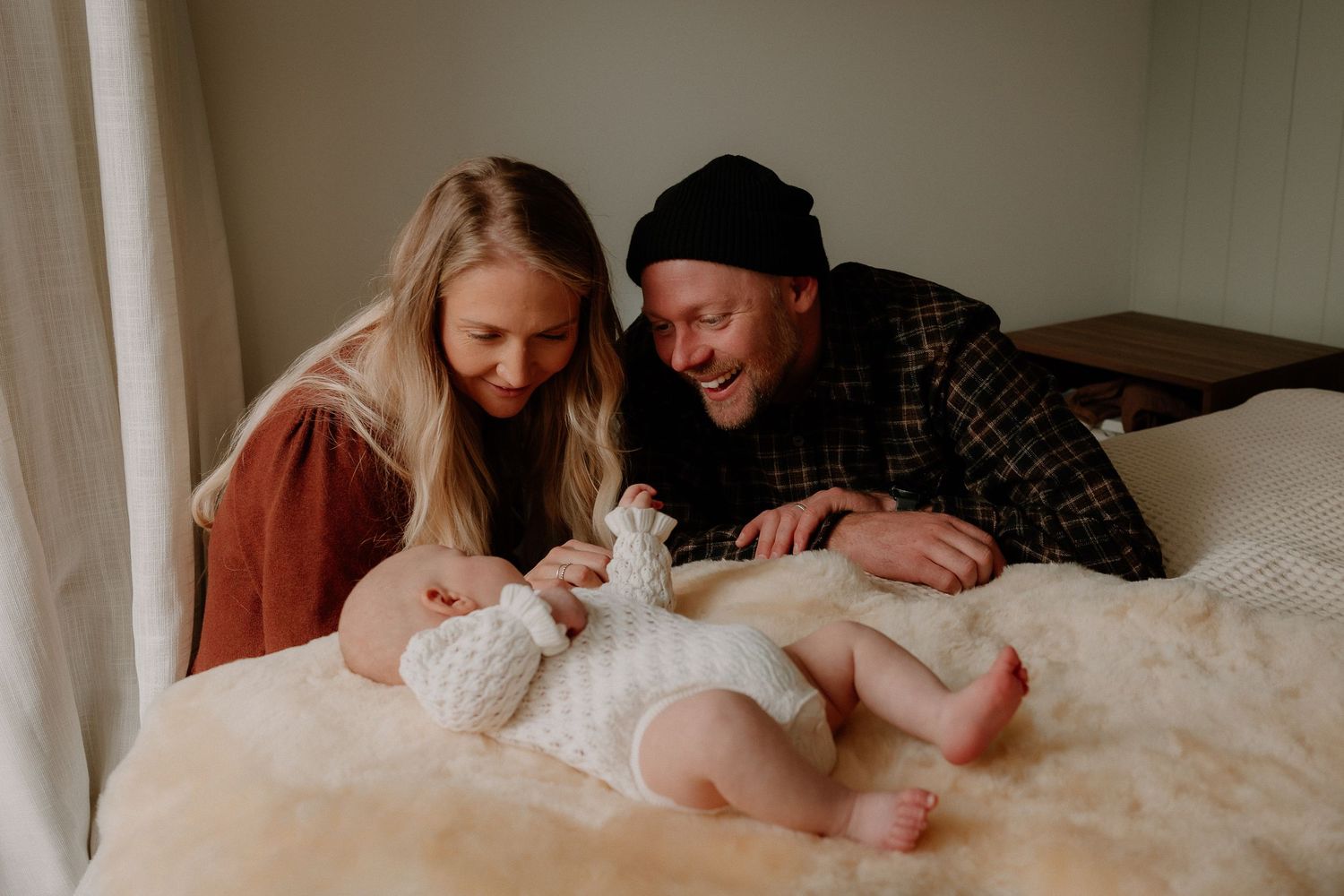 Happy parents lying on a white bed smiling at their newborn baby dressed in a white knit outfit.
