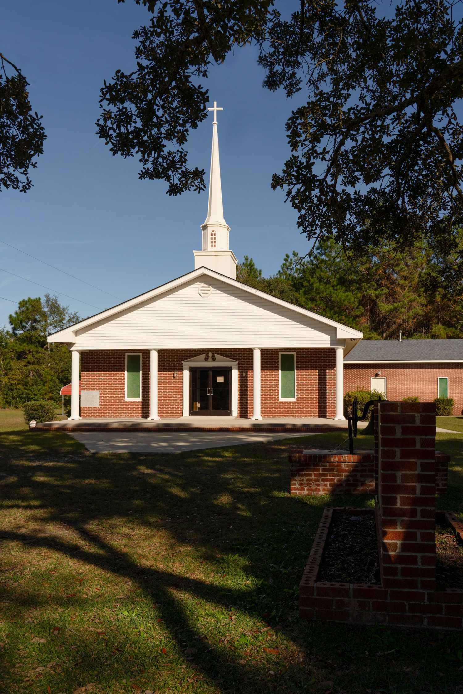 A classic red brick church with white siding and tall steeple topped with a cross, surrounded by green trees under a blue sky.