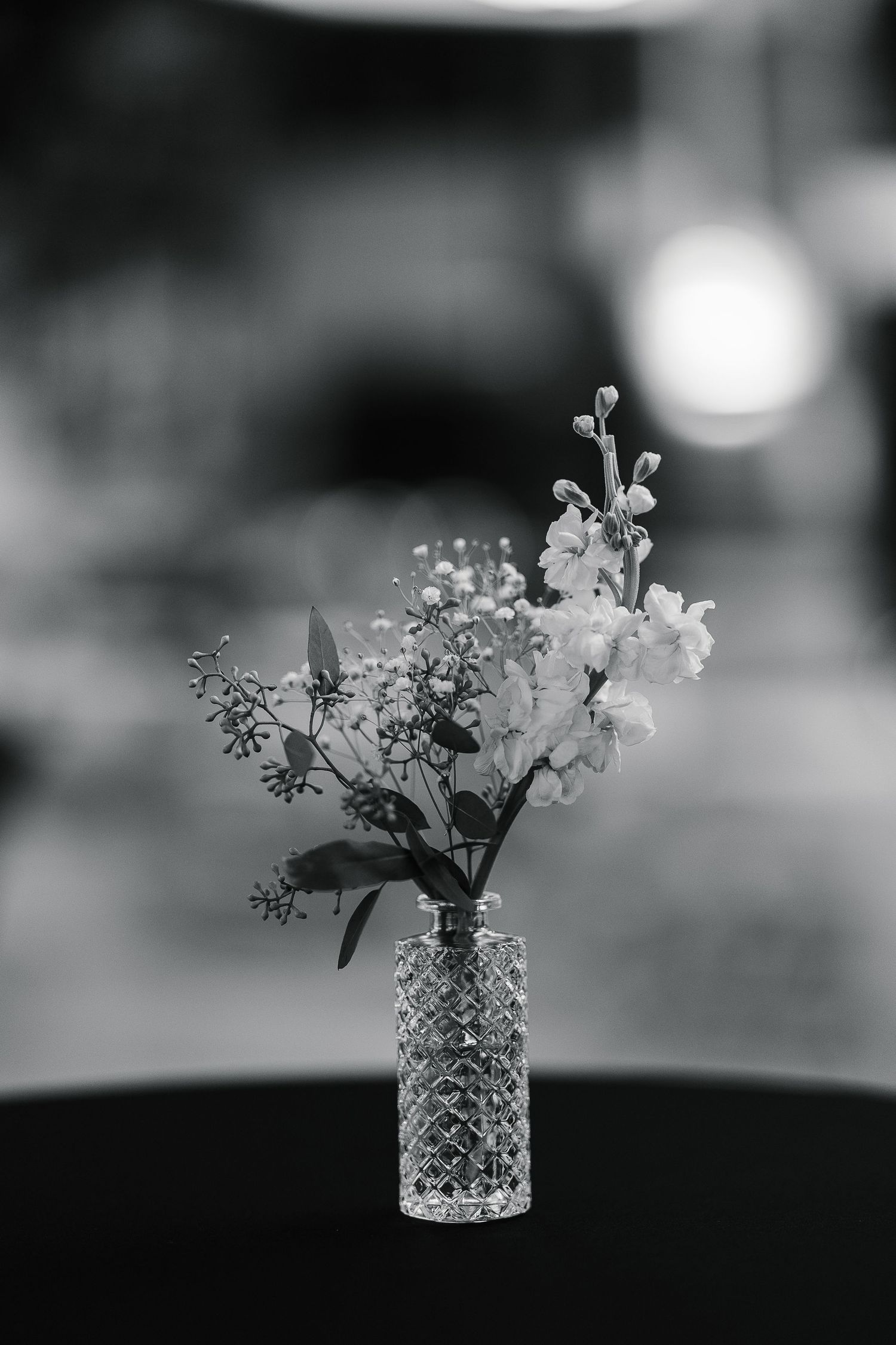 Black and white photo of delicate wildflowers in a crystal vase on a dark table with blurred bokeh background.