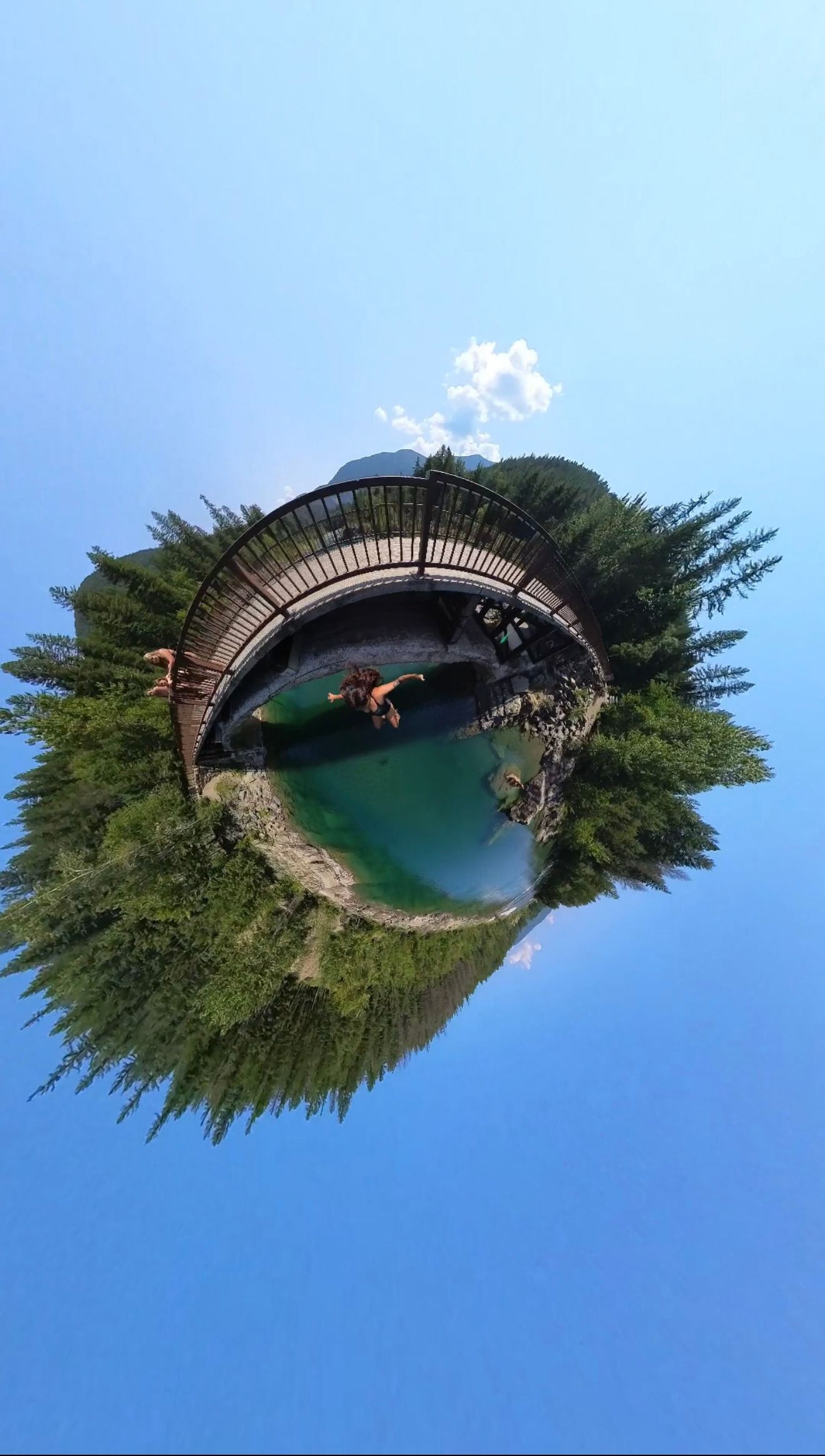 Creative tiny planet 360 photo of a wooden bridge over turquoise lake surrounded by green trees.